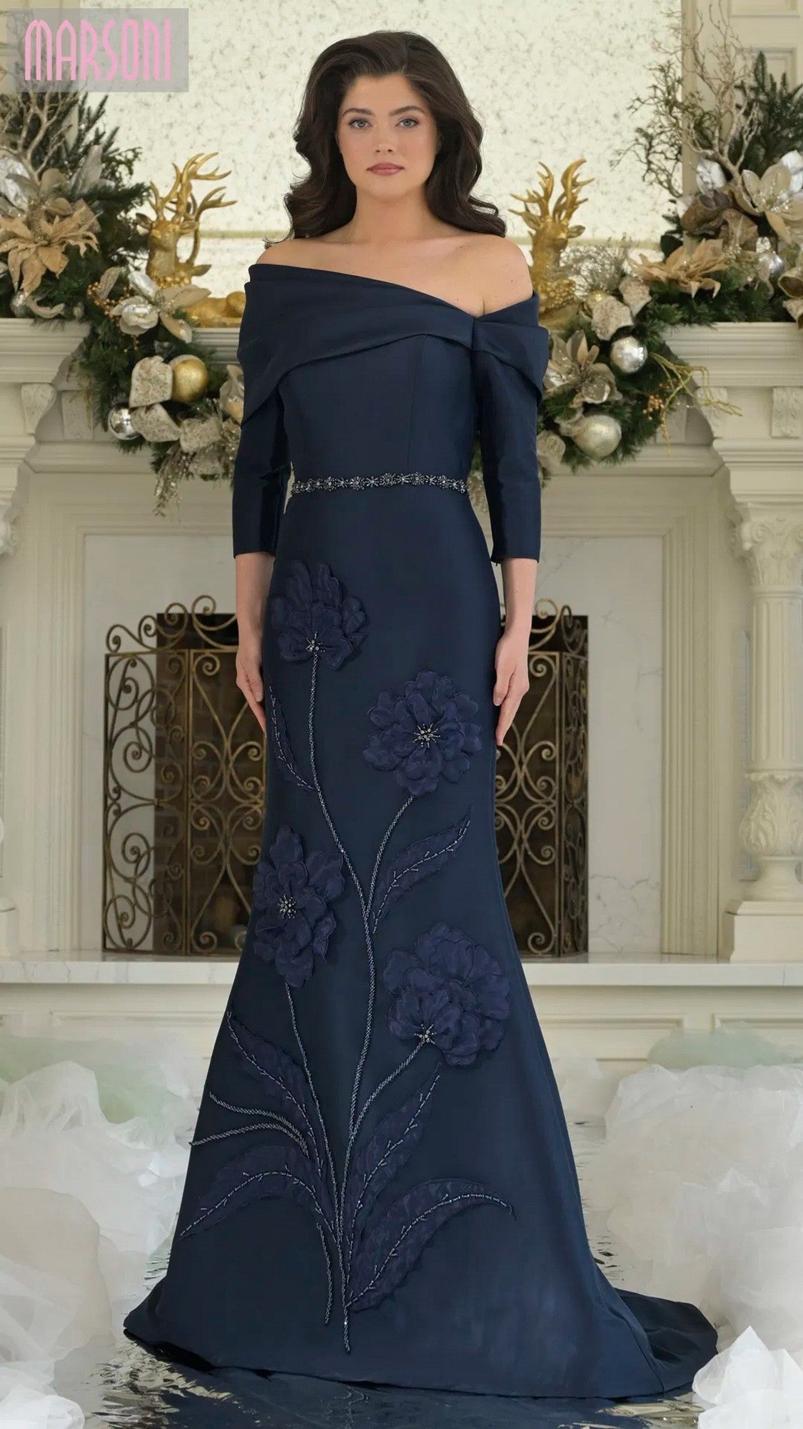A woman stands indoors in front of a decorated white fireplace adorned with holiday garlands, wearing the navy Marsoni MV1374 long formal dress with large floral applique and an off-shoulder neckline.